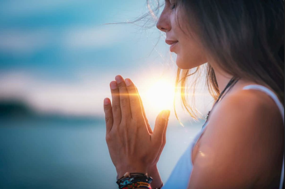 Woman with long hair holding hands together like in prayer outside in front of a sunset